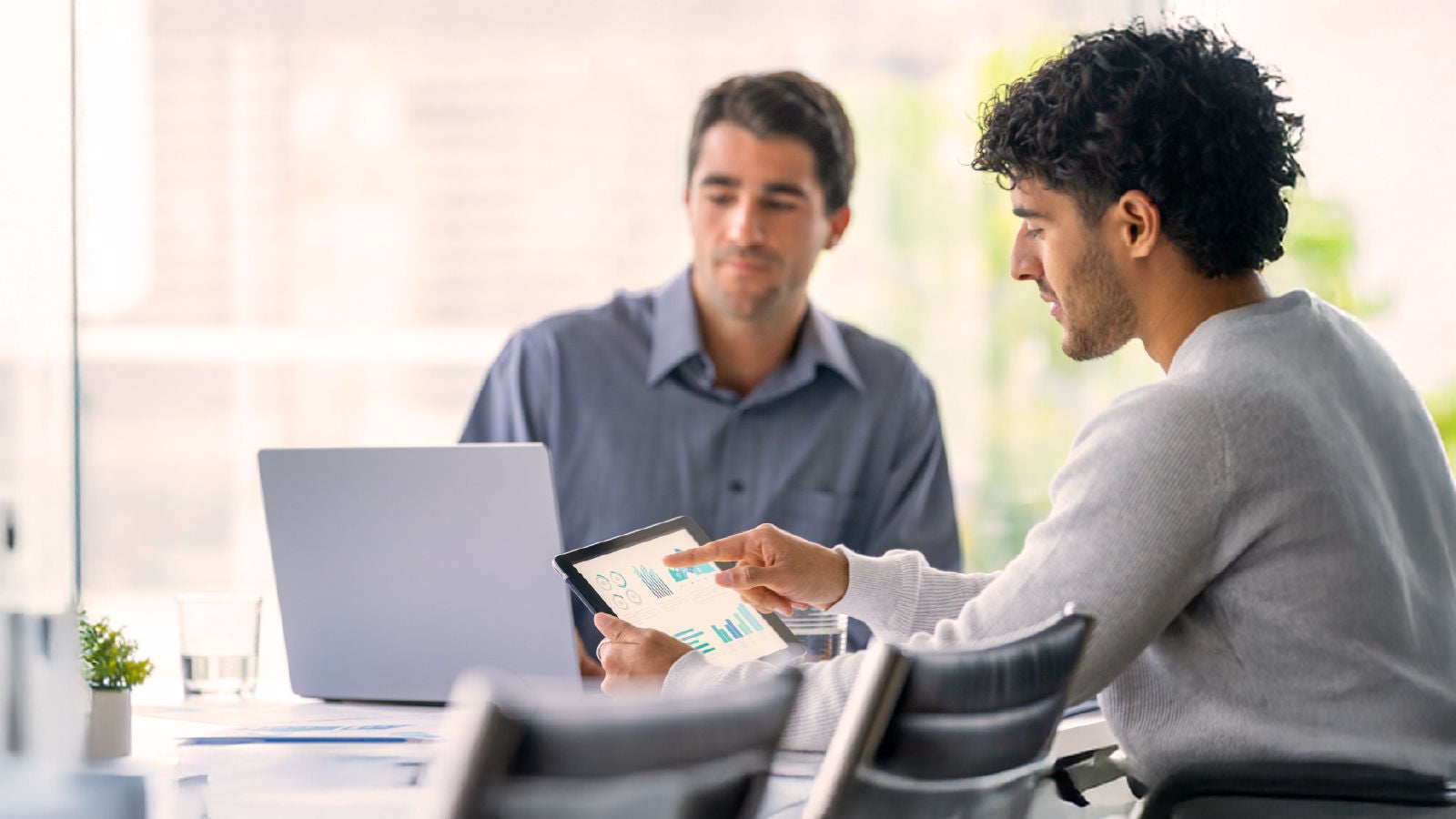 Two professionals collaborating at a table while reviewing charts and data displayed on a tablet.