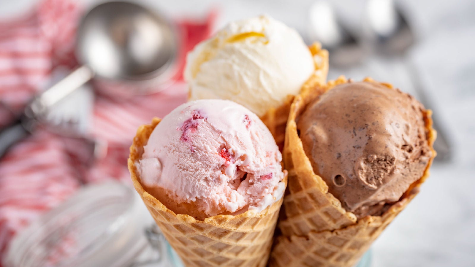 Three scoops of ice cream in waffle cones, including strawberry, vanilla, and chocolate flavors, with an ice cream scoop and kitchen utensils blurred in the background.