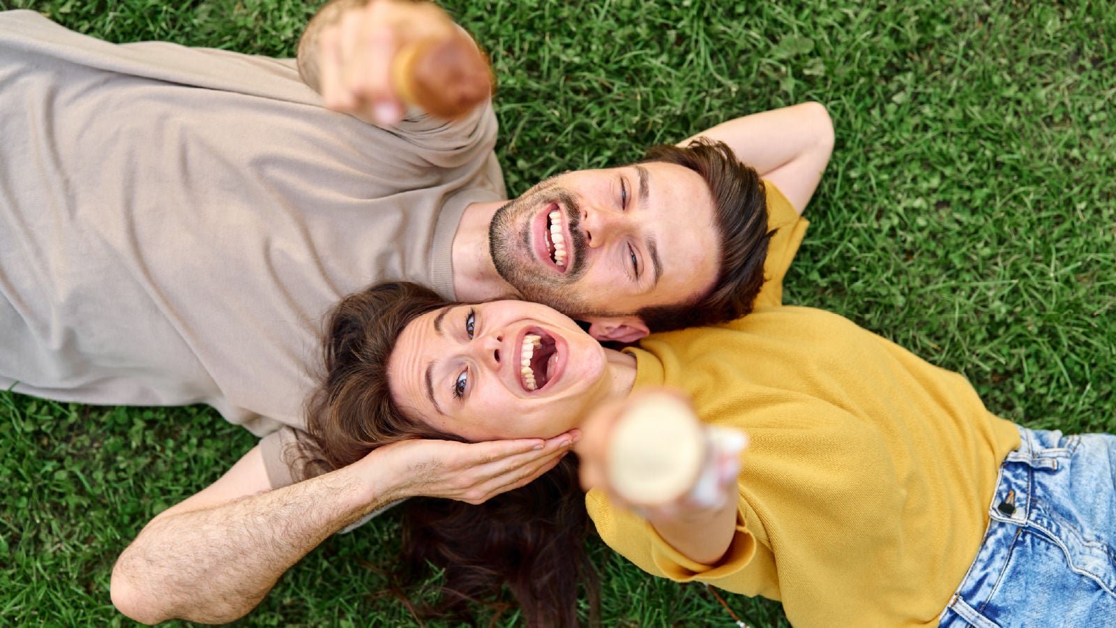 A smiling couple lying on grass outdoors holding ice cream cones while taking a selfie together.