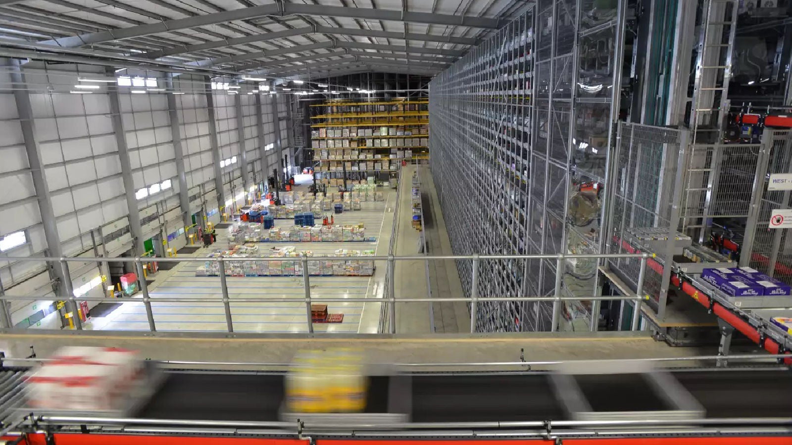A wide view of an Asda warehouse interior, showing organized shelves, conveyor belts, and workers managing inventory and logistics.