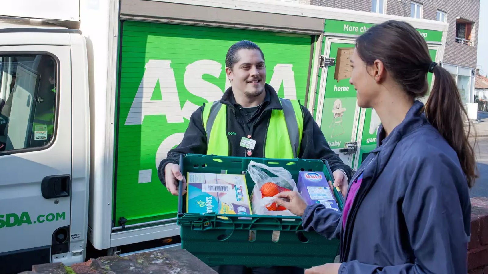 An Asda delivery worker handing a crate of groceries to a customer outside a delivery van with the Asda.com logo in the background.