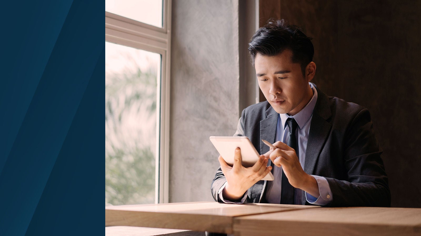 Professional in a suit using a digital tablet at a wooden table by a window, with natural light coming in and a blurred leafy background outside.