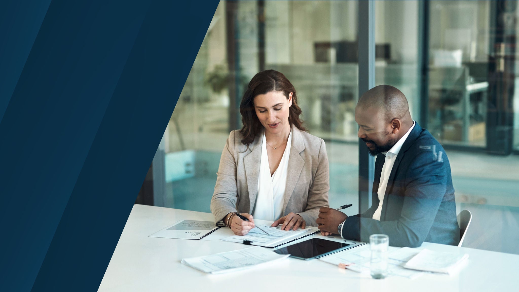 A woman and a man in business attire seated at a modern office table, reviewing financial documents together with notebooks, a tablet, and papers in front of them.