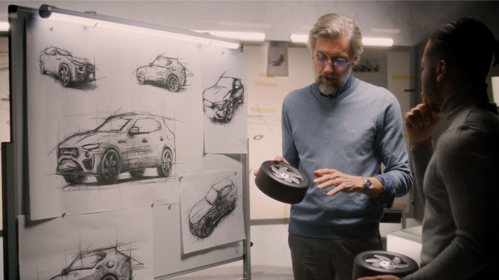 Two automotive design professionals review vehicle sketches and a wheel model beside a display board.