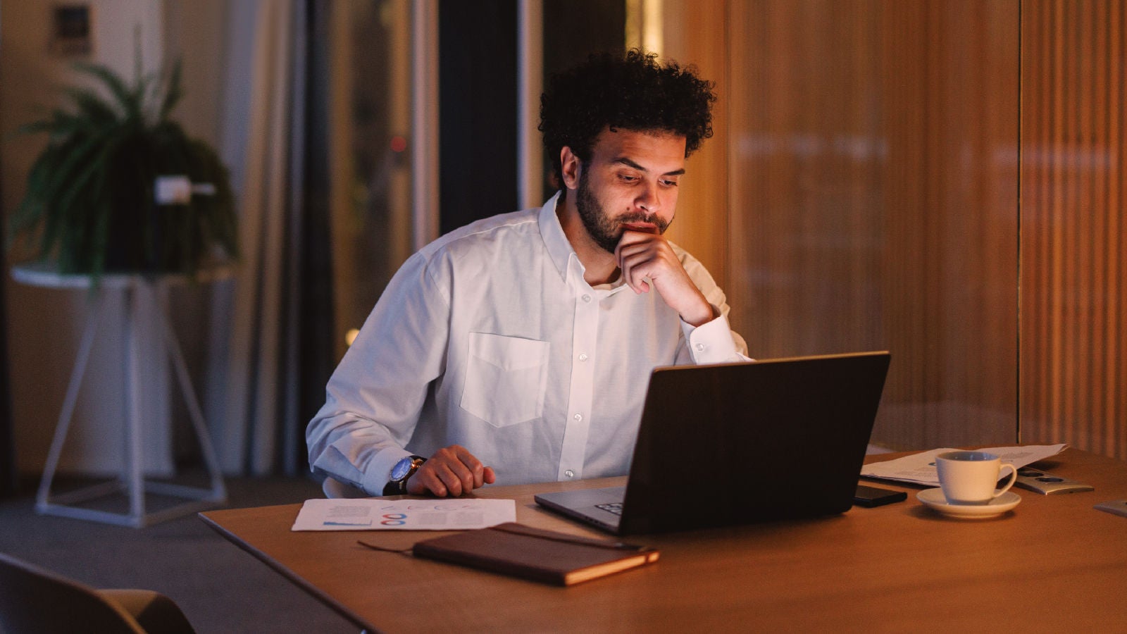 A man in a white shirt working at a desk in a dimly lit office, focused on a laptop screen. Papers, a notebook, and a cup of coffee are visible on the table.