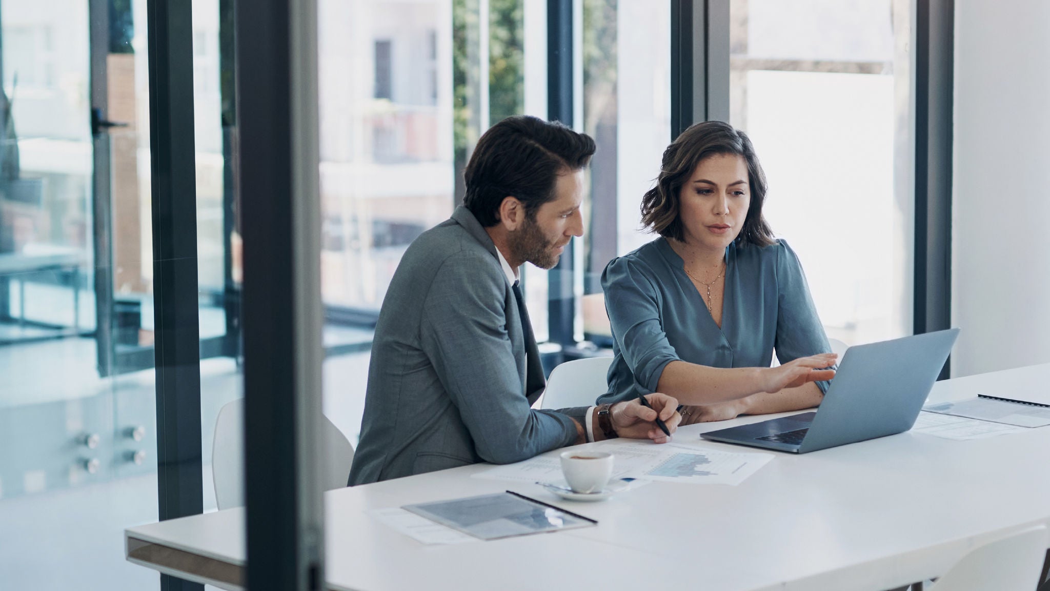 Two coworkers sitting at a table in an office talking over a laptop together