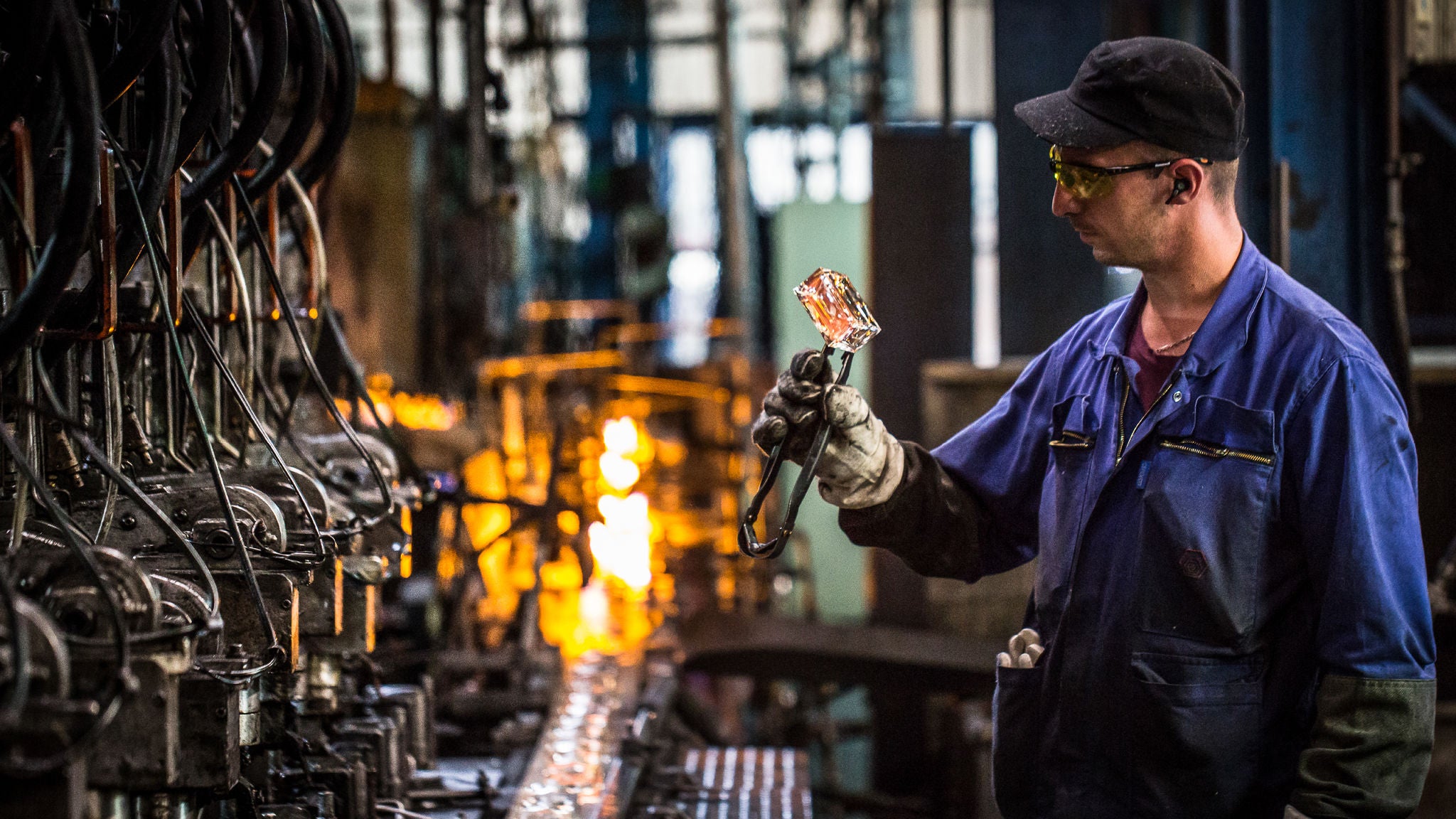 A factory worker in safety gear inspects a glowing glass component using tongs, surrounded by industrial machinery and furnaces in operation.