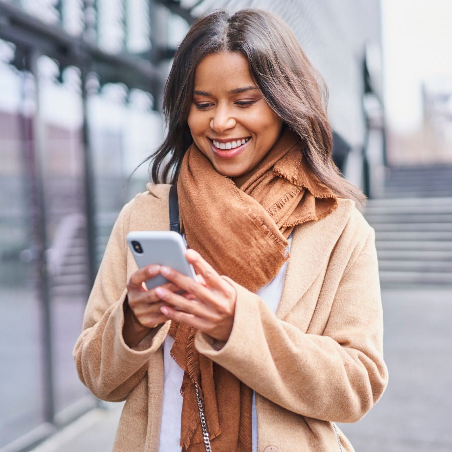 A woman in a brown coat and scarf smiles while using her phone, exuding warmth and joy in a casual setting.