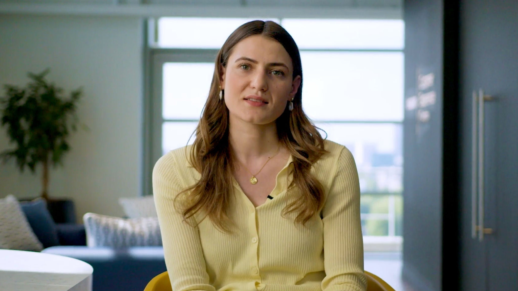 A woman wearing a yellow cardigan sits indoors, facing the camera, with a blurred office background and natural light coming through a window.