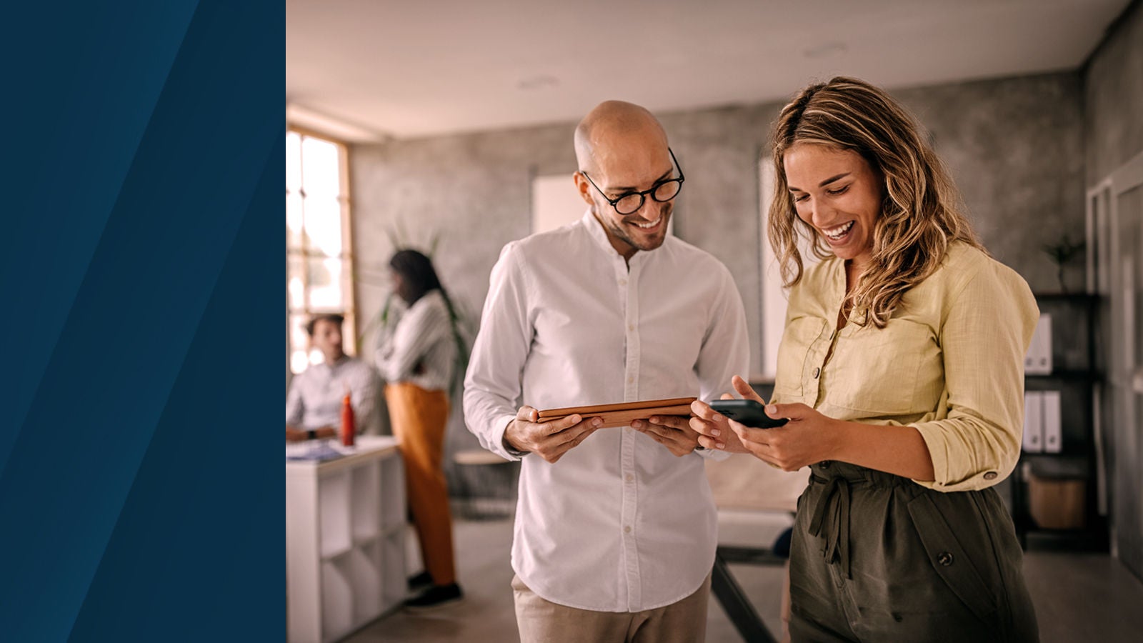 Two colleagues smiling and looking at a tablet and smartphone in a bright, modern office space, with two other coworkers collaborating in the background.