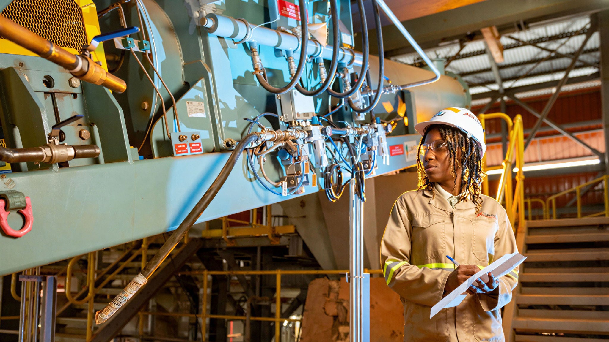 Worker in protective gear inspecting industrial equipment inside a processing facility while holding a clipboard.