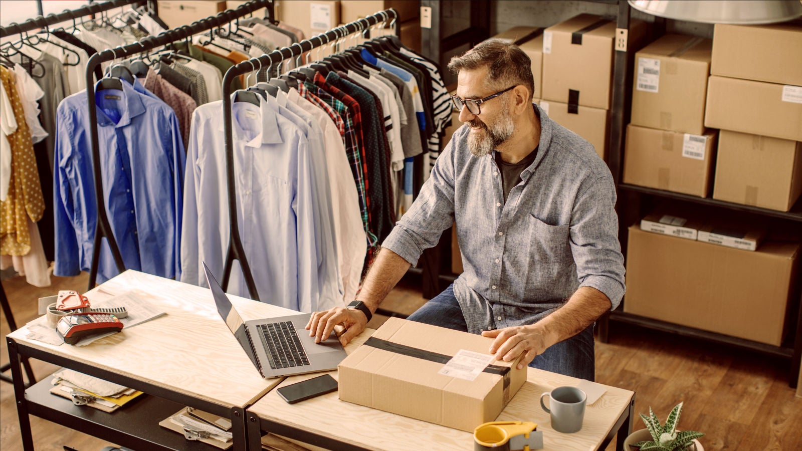 A man working at a desk in a clothing stockroom, surrounded by shipping boxes and racks of shirts, while using a laptop and handling a sealed parcel.