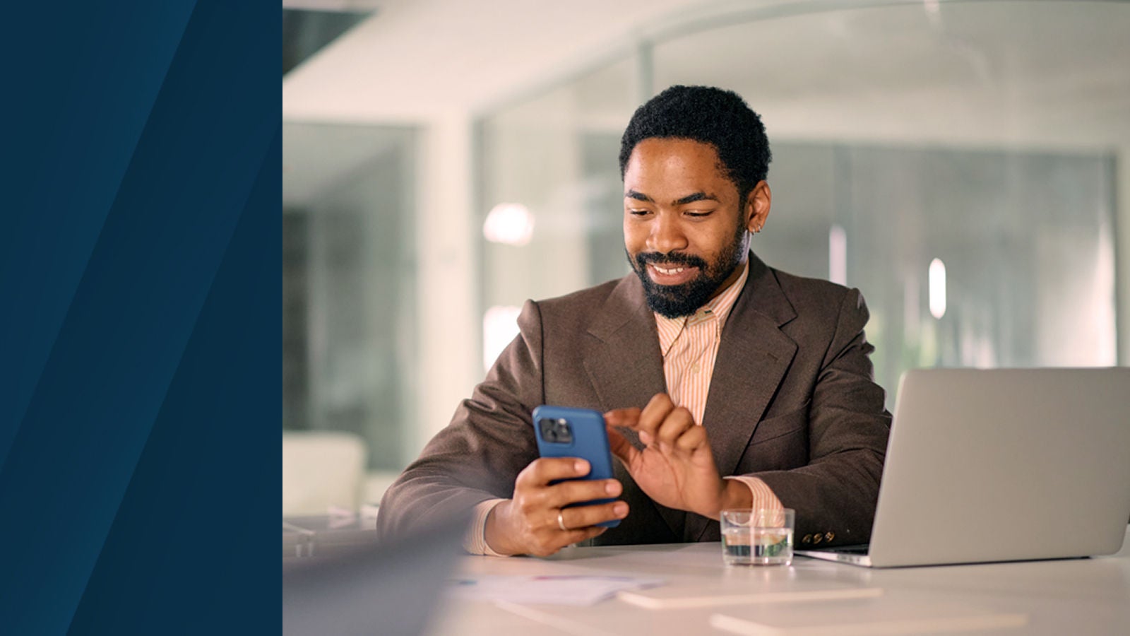Man in a brown suit sitting at a desk with a laptop and glass of water, smiling while using a smartphone.