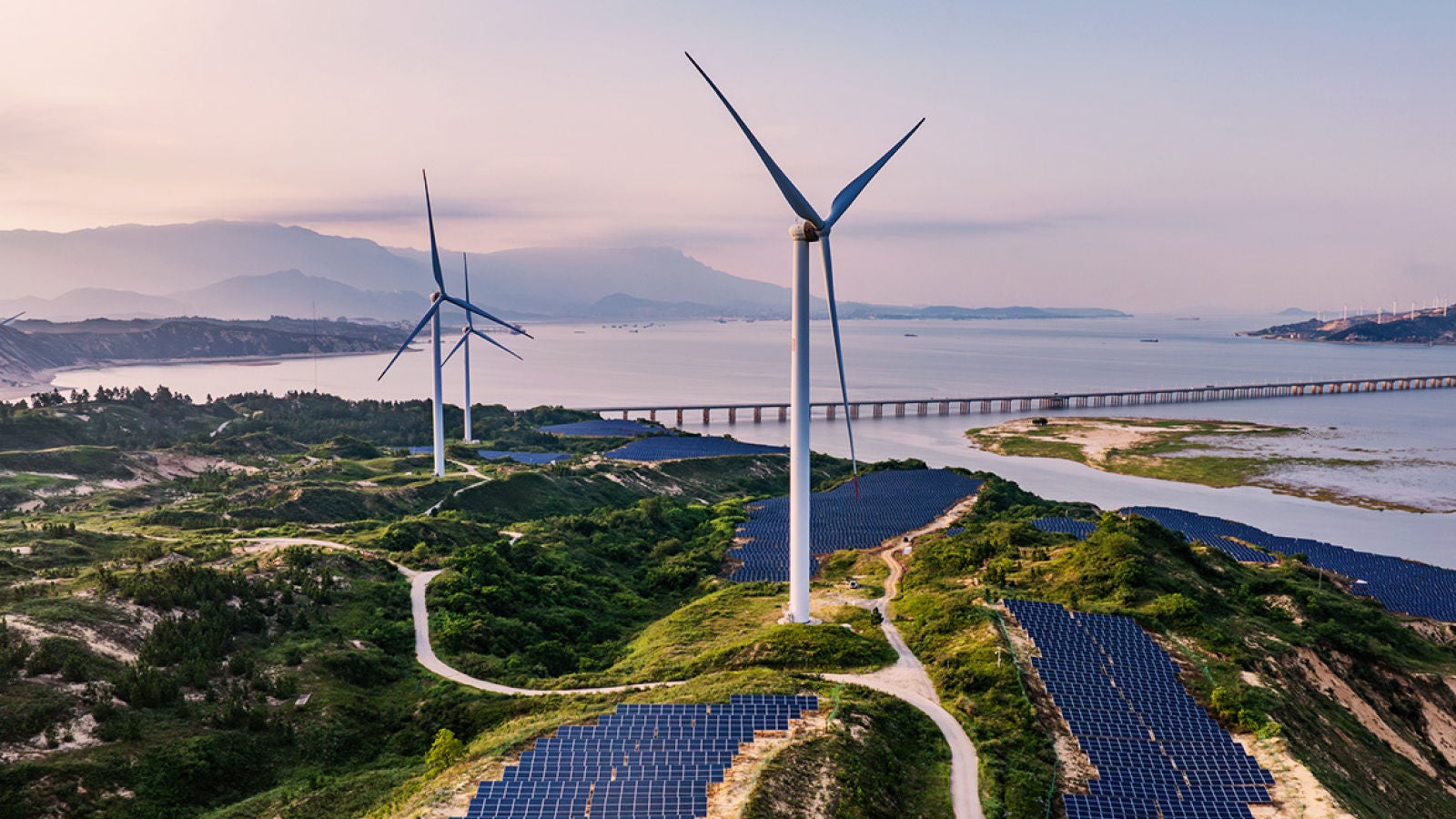 Wind turbines and solar panels across coastal hills near a bridge.