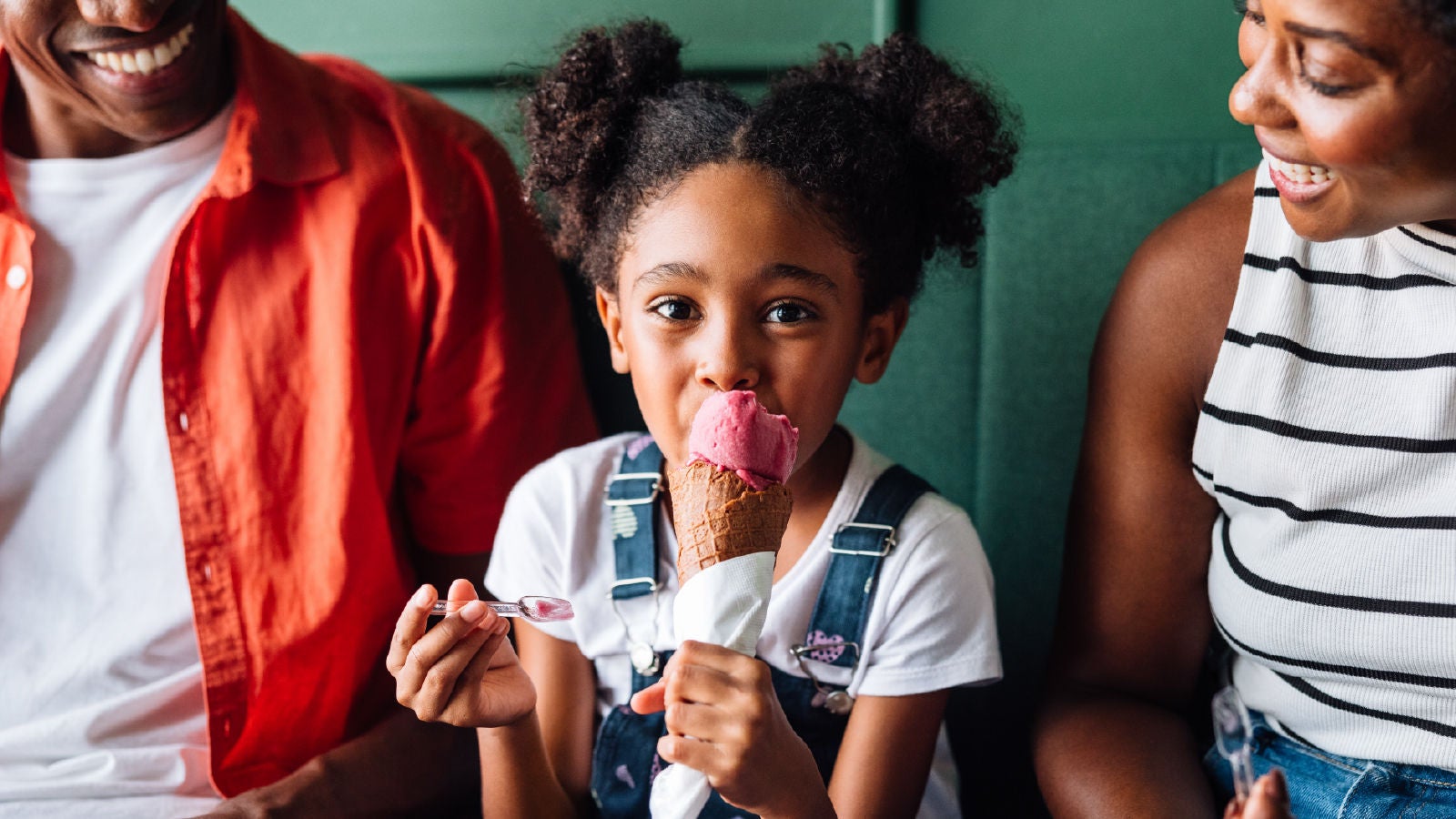 A young girl eating a pink ice cream cone while sitting between two adults, smiling and enjoying ice cream together.