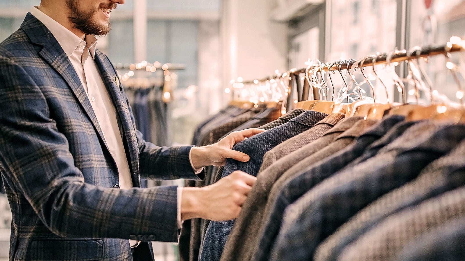 Close-up of a man in a blue plaid blazer examining coats on hangers in a well-lit retail clothing store decorated with warm string lights.