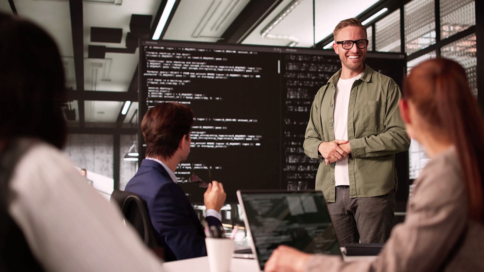 A business professional presents to colleagues in a meeting room with code displayed on large screens.