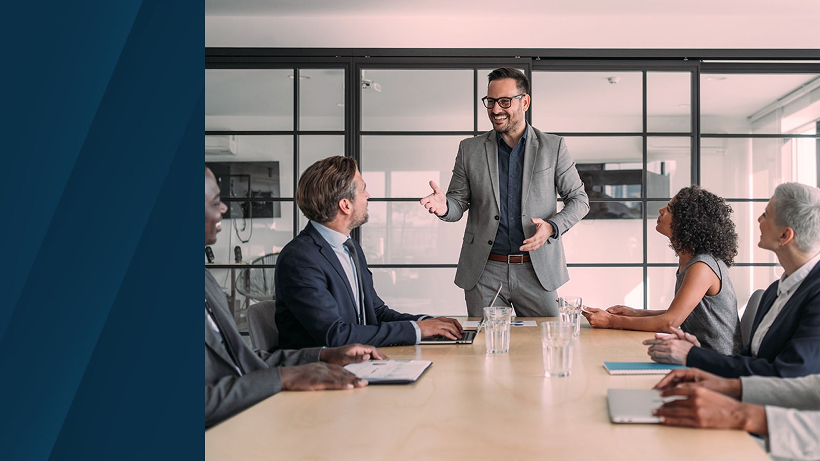 A man in a grey suit stands at the head of a conference table, speaking to a group of colleagues seated and listening attentively in a modern glass-walled meeting room.