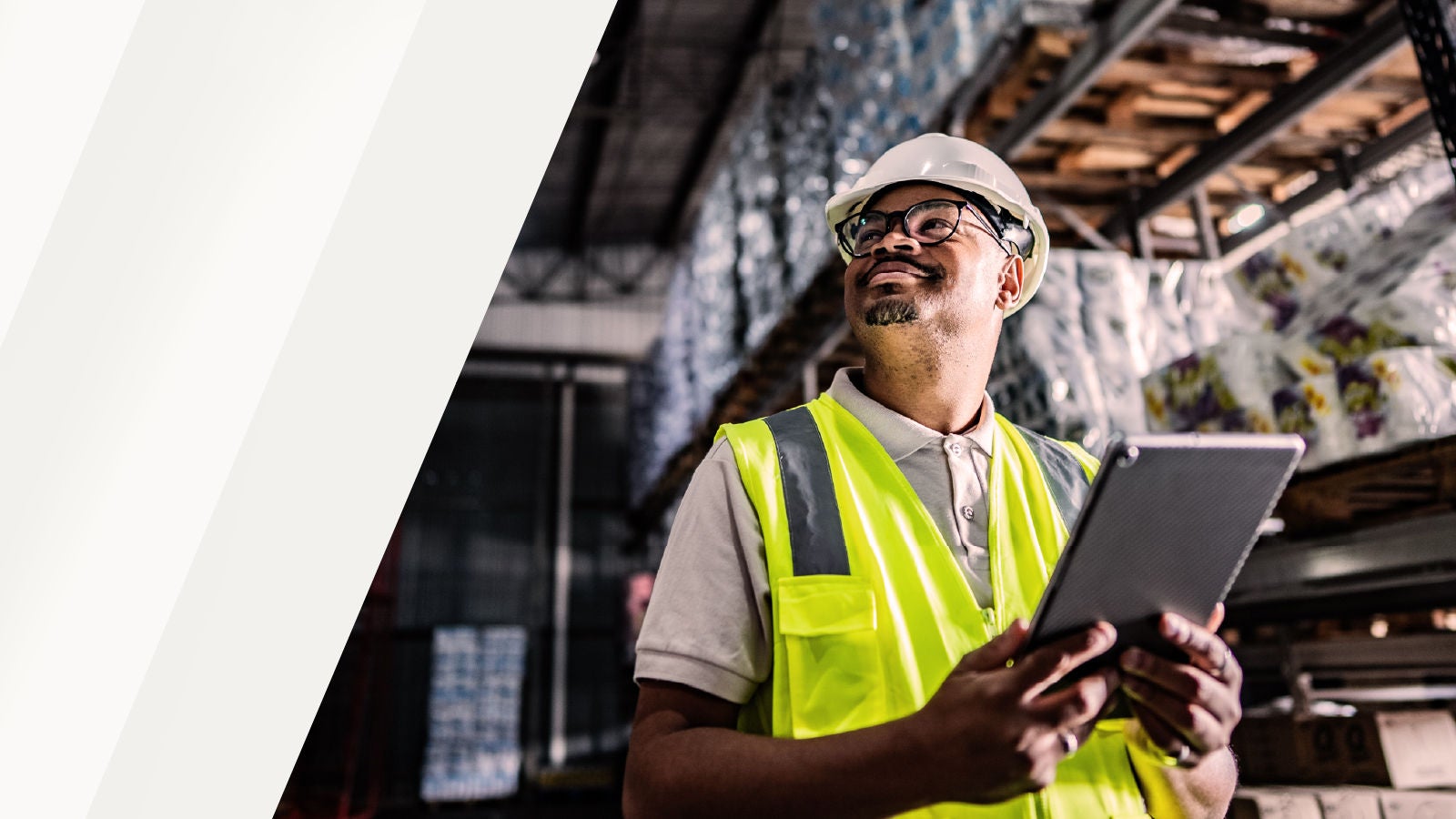 Employé d’entrepôt portant un gilet de sécurité et un casque, tenant une tablette dans une allée de centre de distribution avec des palettes de marchandises en arrière-plan.