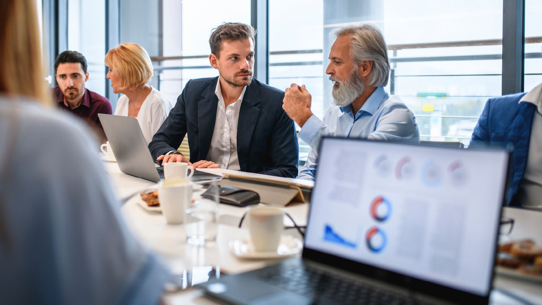 Business professionals in a meeting room engaged in discussion, with charts displayed on a laptop in the foreground.
