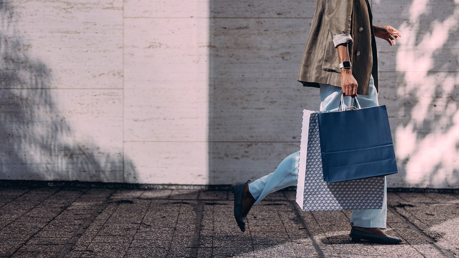 A person walking along a textured sidewalk holding two patterned shopping bags. The individual is dressed in light blue trousers, a khaki blazer, and loafers, with a smartwatch visible on their wrist. Soft shadows from nearby trees fall across a pale stone wall in the background.