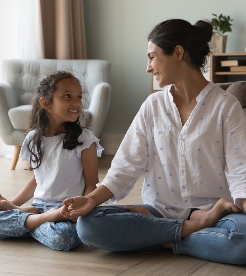 Mother and daughter sitting cross-legged on the floor, smiling at each other in a cozy living room.