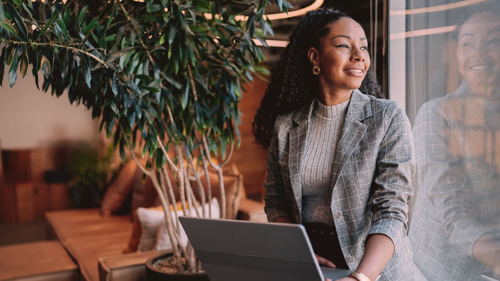 A woman in a plaid blazer sits by a window with a laptop, smiling as she looks outside. The setting includes a potted tree and cozy seating in a warmly lit room.