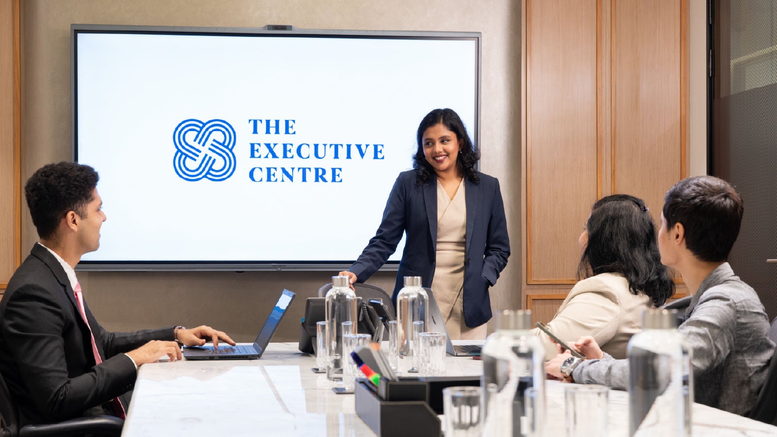 Business professional presents to three colleagues in a meeting room with The Executive Centre logo displayed on a screen.