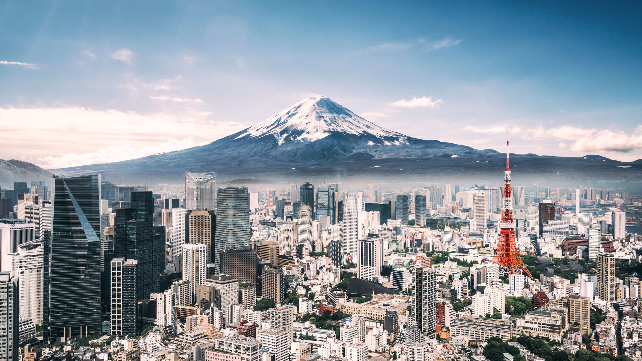 A wide cityscape of Tokyo with Mount Fuji in the background, dense urban buildings in the foreground, and a clear blue sky.
