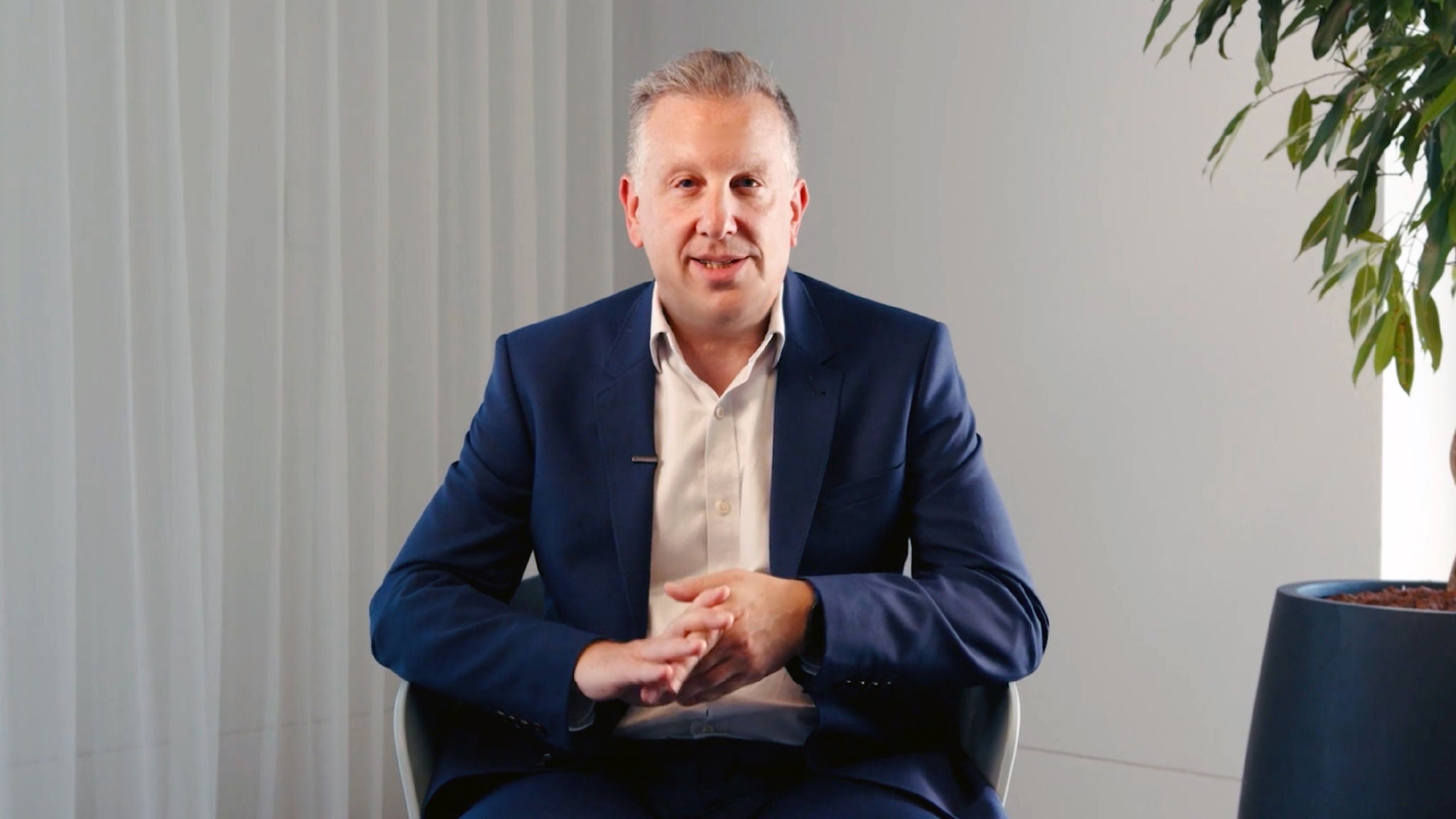 A middle-aged man in a navy suit sits indoors, speaking directly to camera with hands clasped, in a modern office setting with neutral walls and a potted plant.