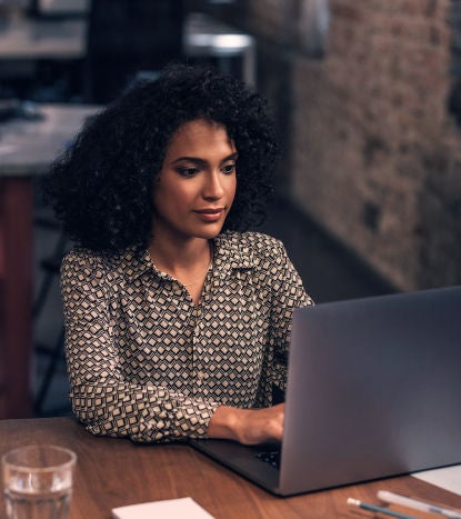 A focused woman working on a laptop in a modern office setting, with a notepad and glass of water on the table.