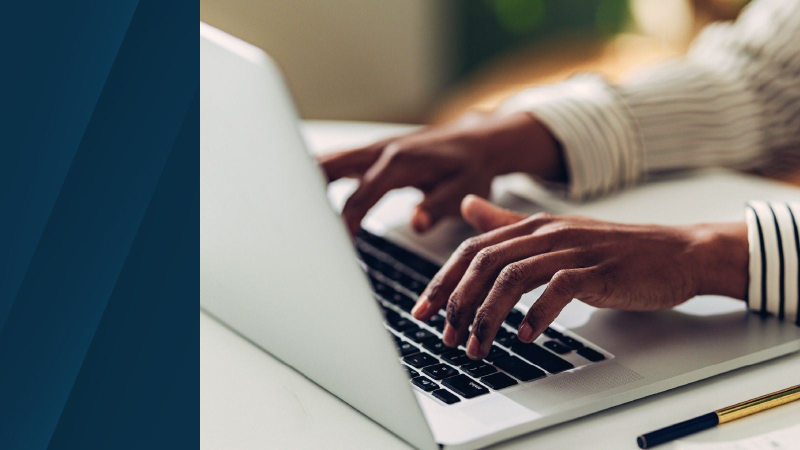 Close-up of hands typing on a laptop keyboard at a desk, with a pen and smartphone nearby, in a brightly lit workspace.