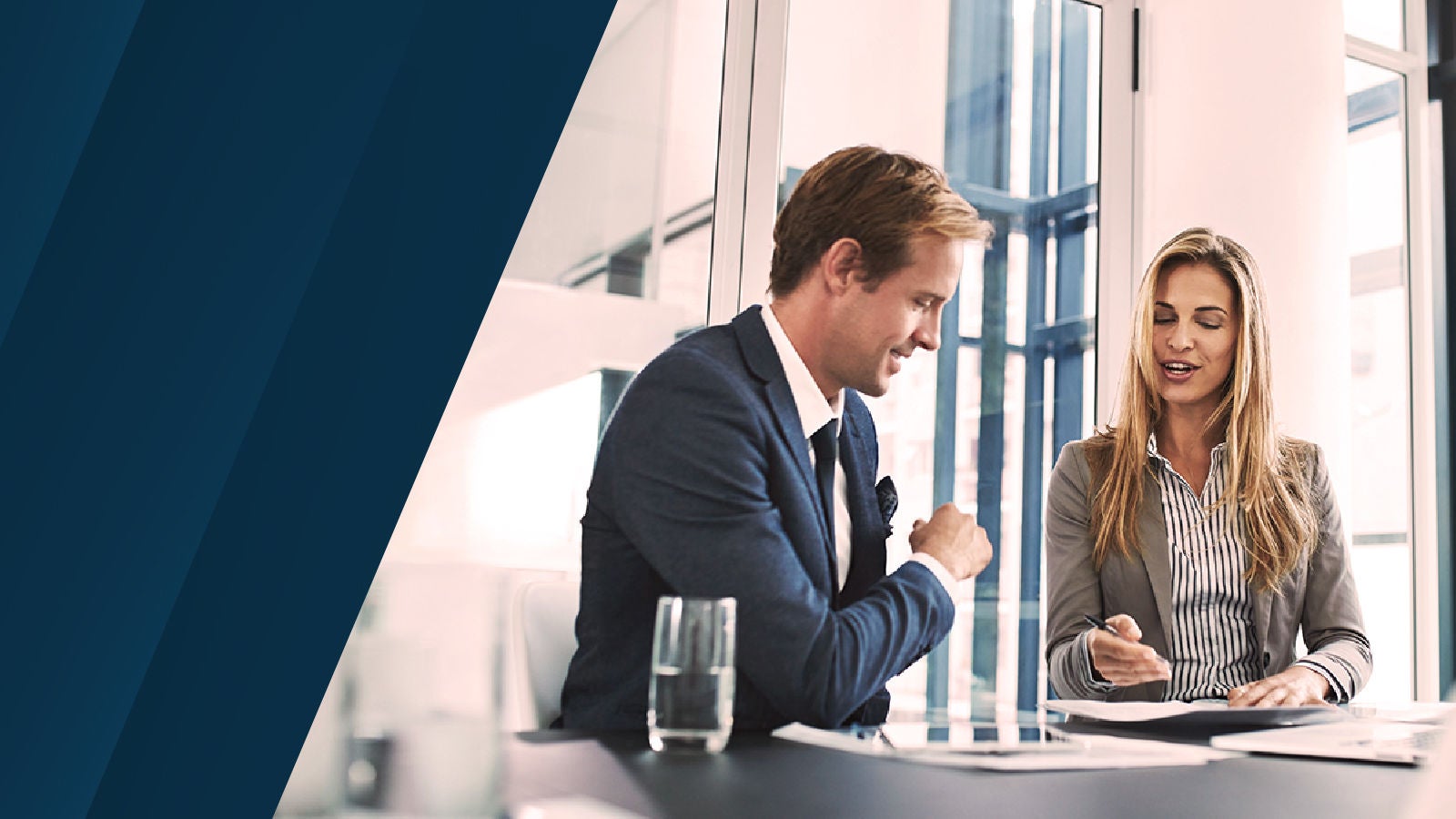The image shows two professionals in conversation across a table in a bright, glass-walled office, suggesting strategic collaboration and planning.