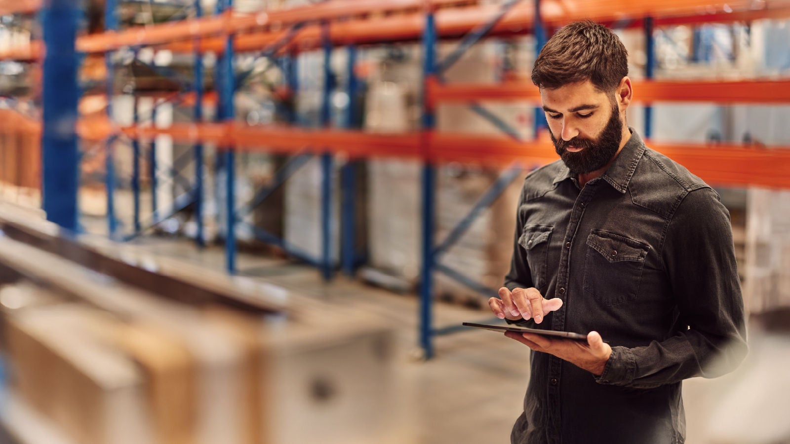 A man with a beard standing in a warehouse aisle, focused on using a tablet, with shelves and boxes in the background.
