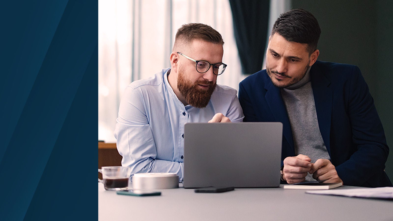 Two professionals sitting at a table, reviewing data on a laptop and discussing work in a well-lit office environment.