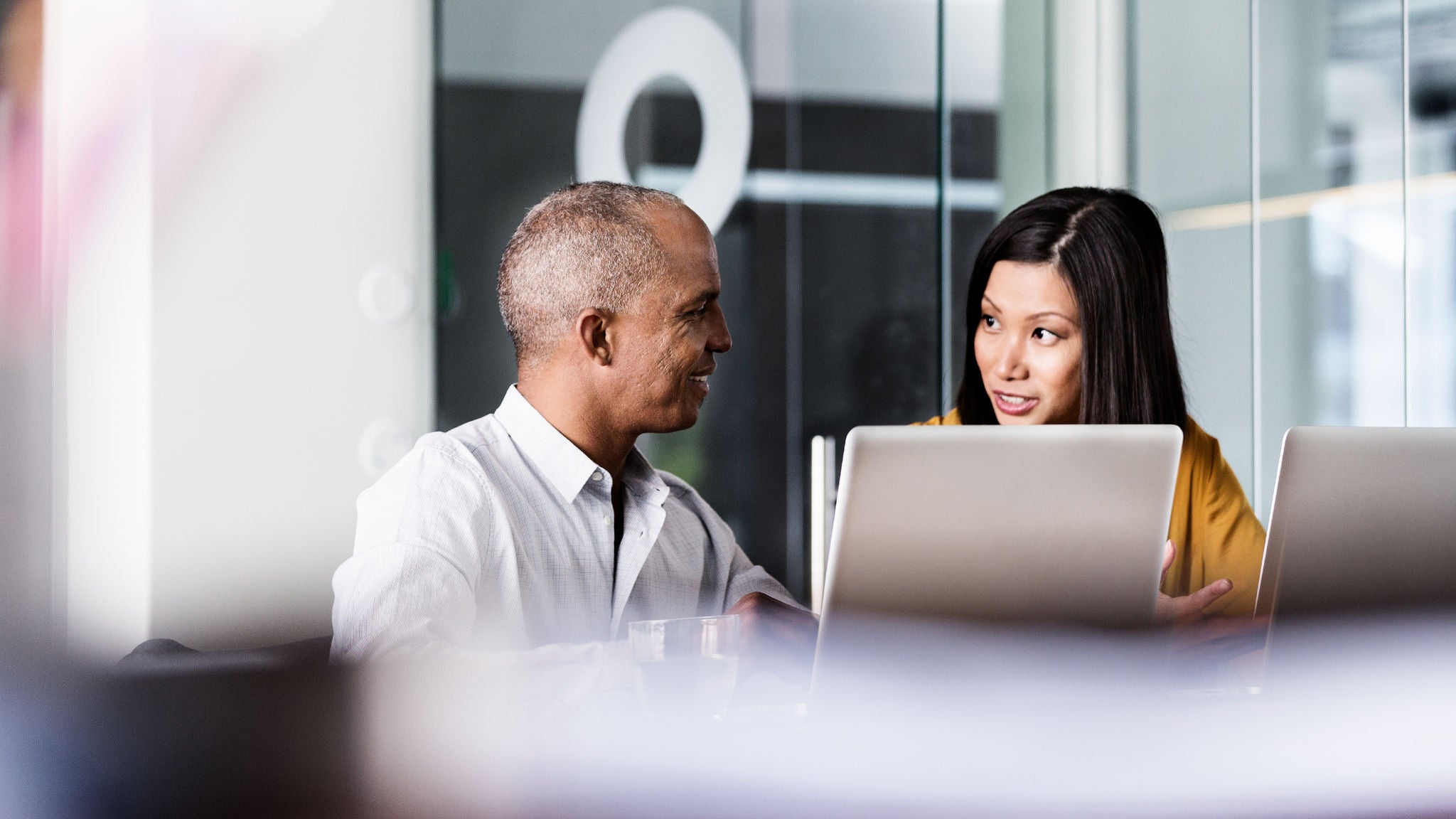 Two professionals seated at a conference table with laptops, engaged in discussion in a bright modern office setting with glass partitions.