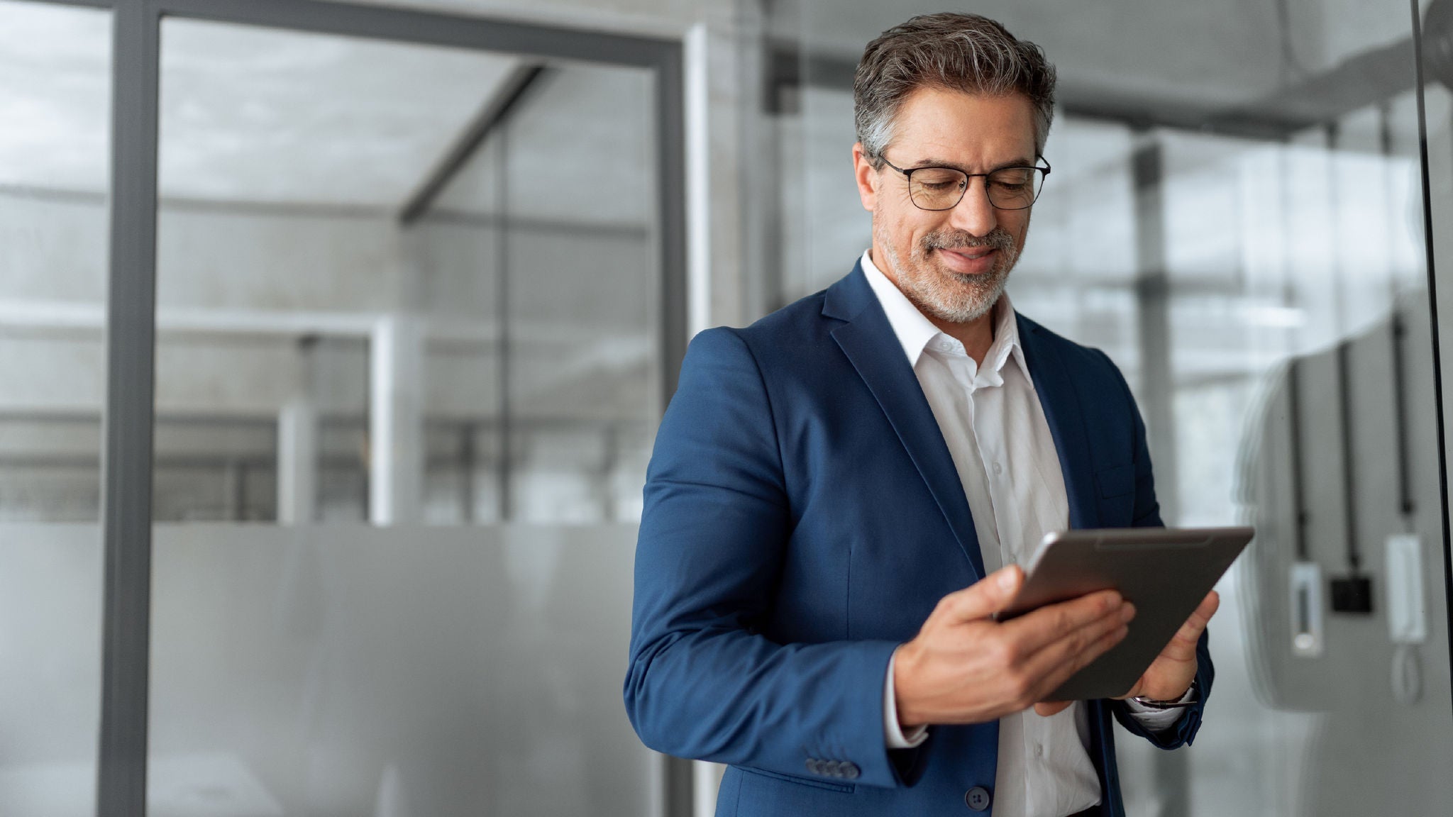 A person in a blue suit jacket and white shirt looking at a digital tablet with a slight smile, standing near glass panels in a bright, professional office environment.