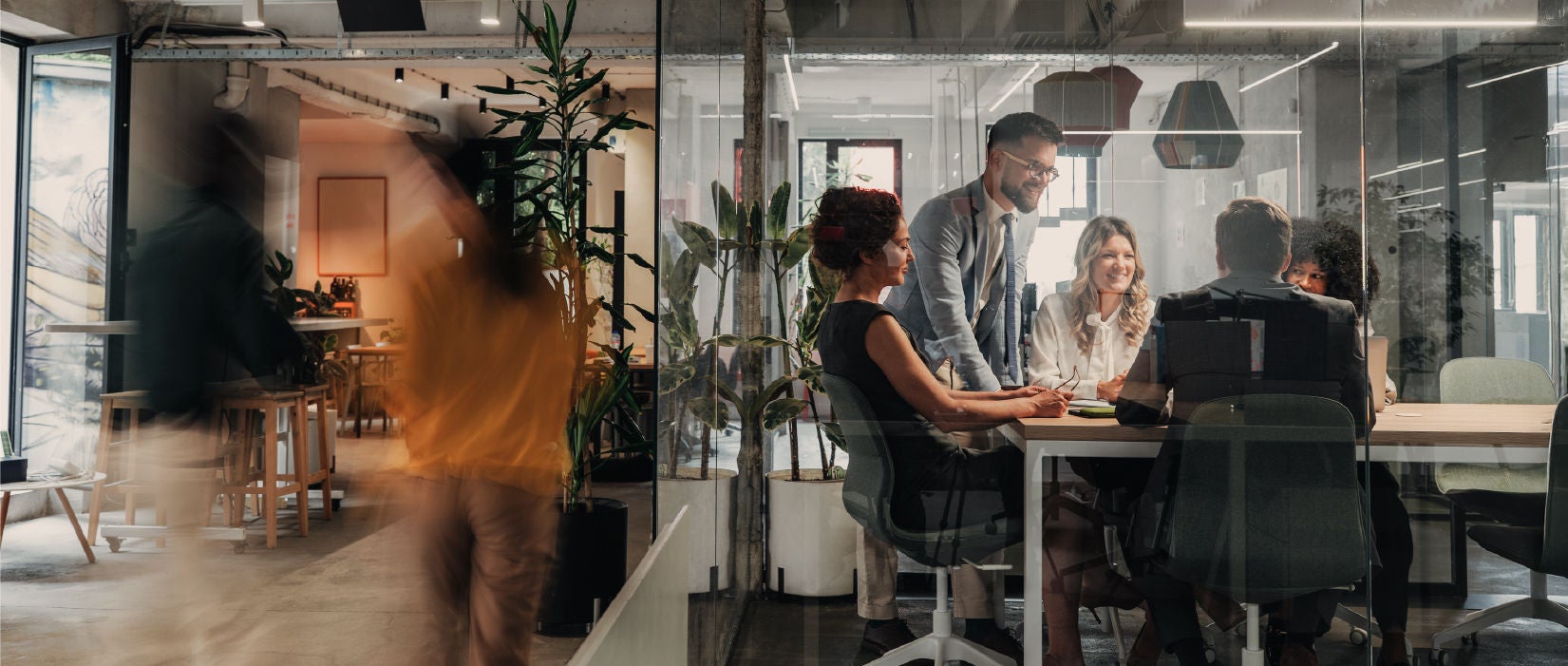 A group of professionals collaborating in a modern glass-walled meeting room, with blurred figures walking by, representing teamwork and a dynamic work environment.