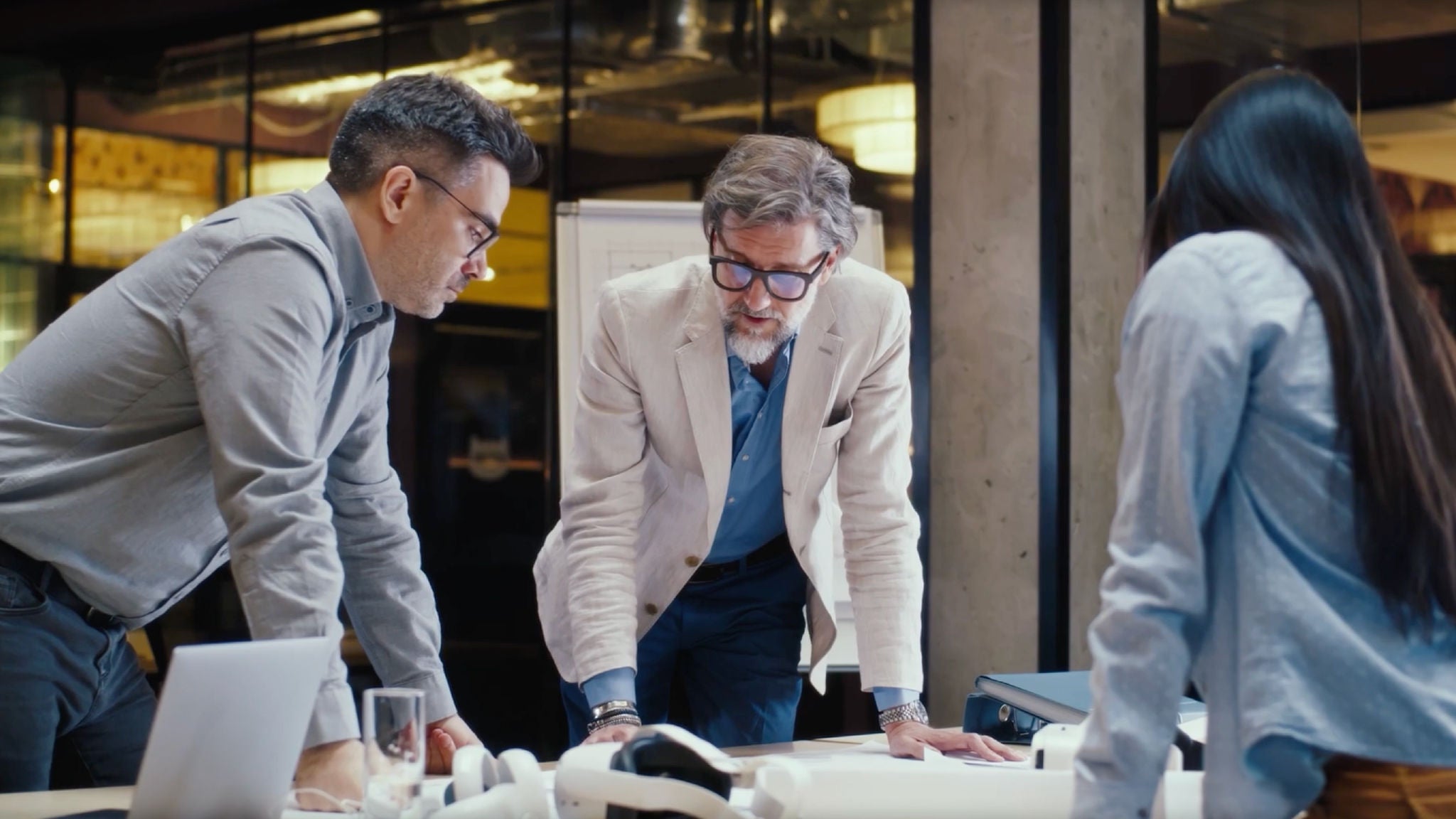 Three professionals lean over a table reviewing documents together in a modern office, with laptops, papers, and a whiteboard visible in the background.