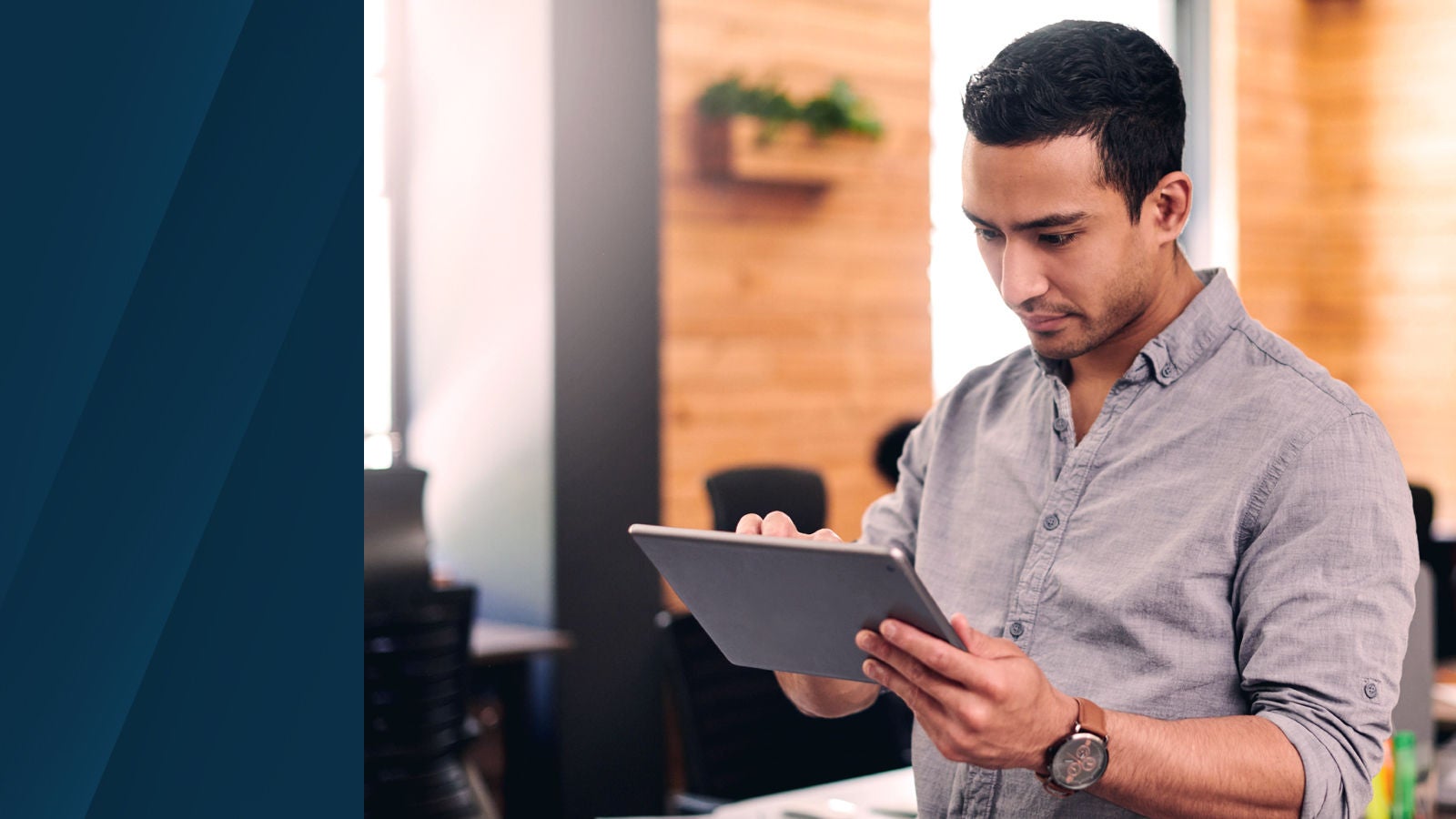 A professional in a gray button-up shirt using a tablet in a modern office setting with wooden accents and greenery in the background.