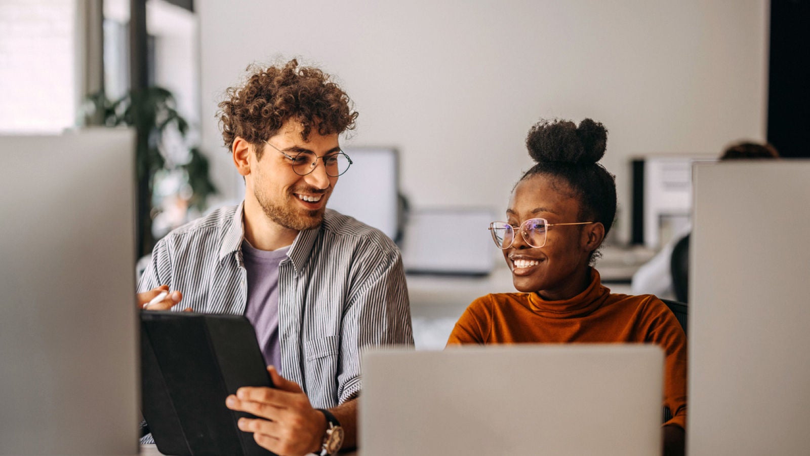 Two colleagues seated at desks, smiling while discussing work. The man holds a tablet while the woman looks on, with laptops visible in front of them.