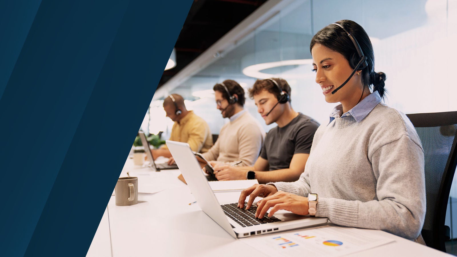 Customer service representatives wearing headsets work on laptops in an office