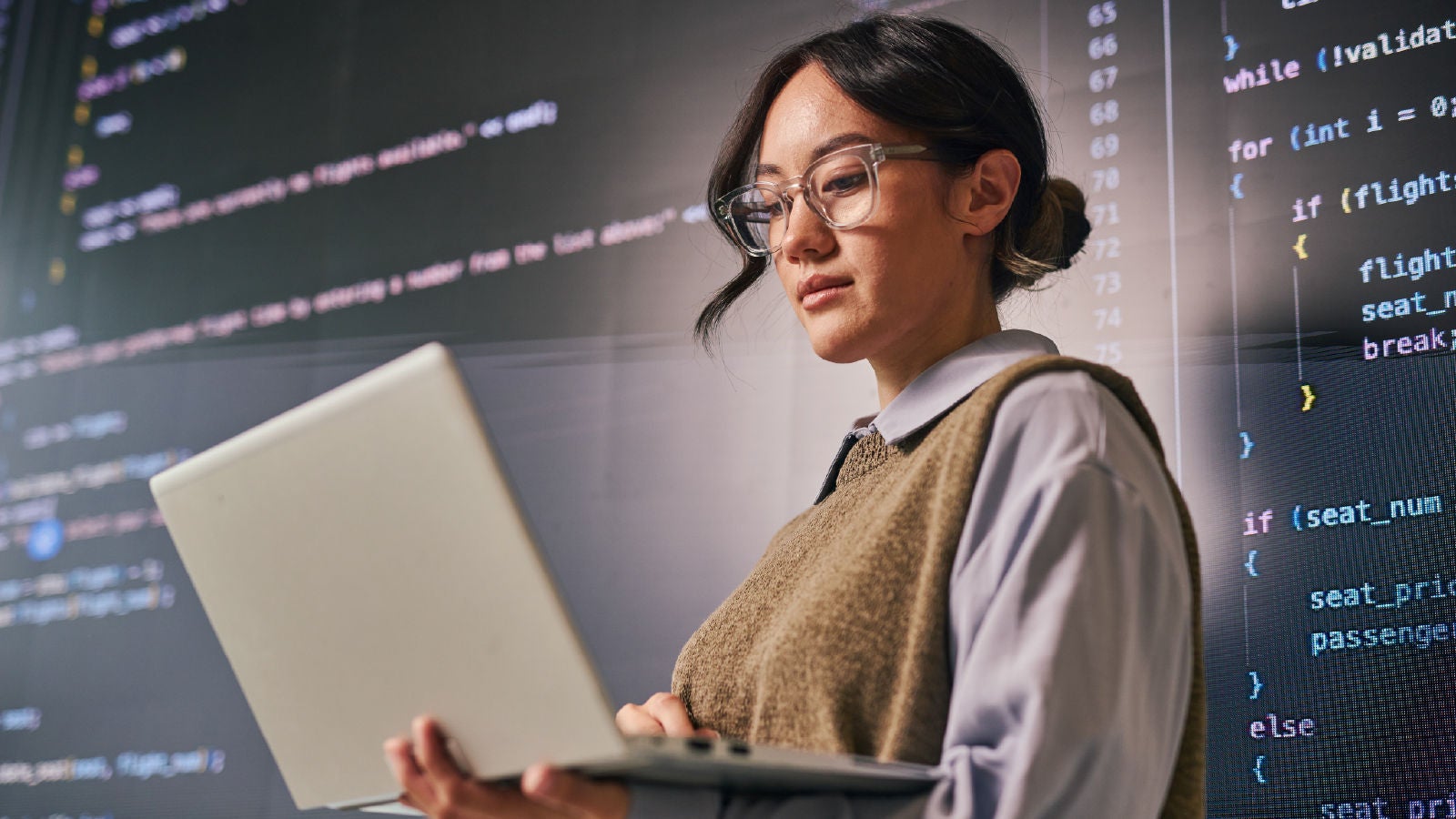 A professional holding a laptop while standing in front of a large screen displaying lines of code.