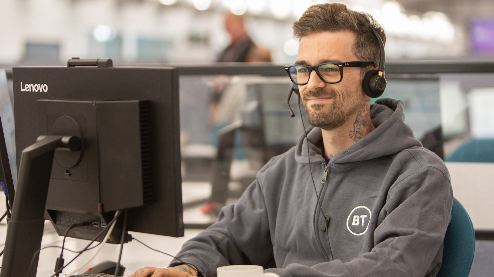 BT employee wearing a headset and glasses working at a desktop computer in an office environment, with the BT logo visible on their sweatshirt.