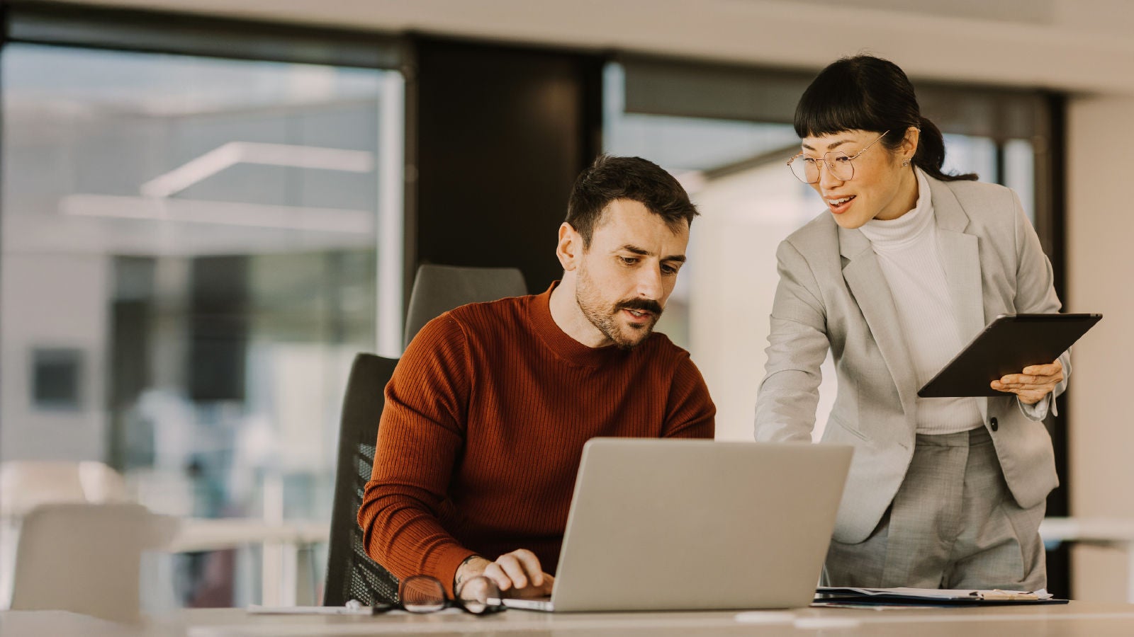 A woman in a light gray suit and a man in a red sweater working together at a laptop in a modern office space, both focused on the screen.