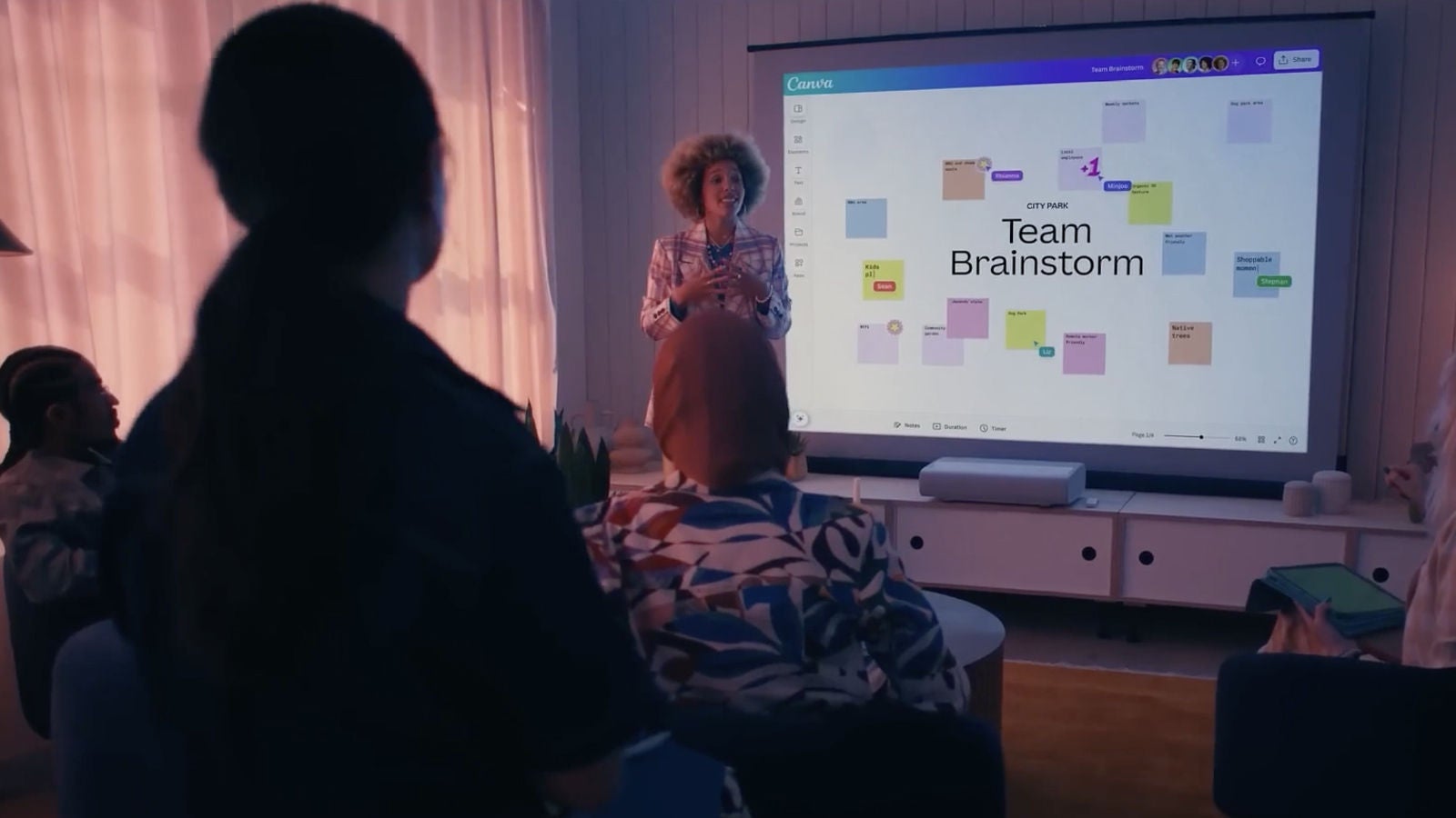 A group gathered in a dimly lit room watching a presenter speak in front of a large screen showing a Canva team brainstorm board.