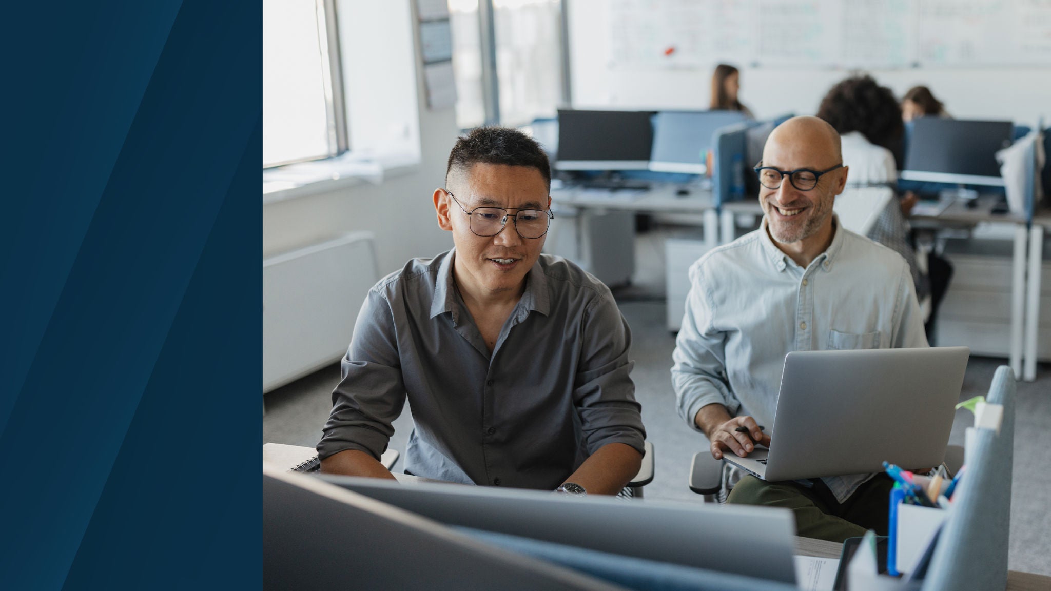 Two colleagues sitting in a modern office, one using a laptop and the other focused on a desktop screen, both smiling and engaged in collaborative work.