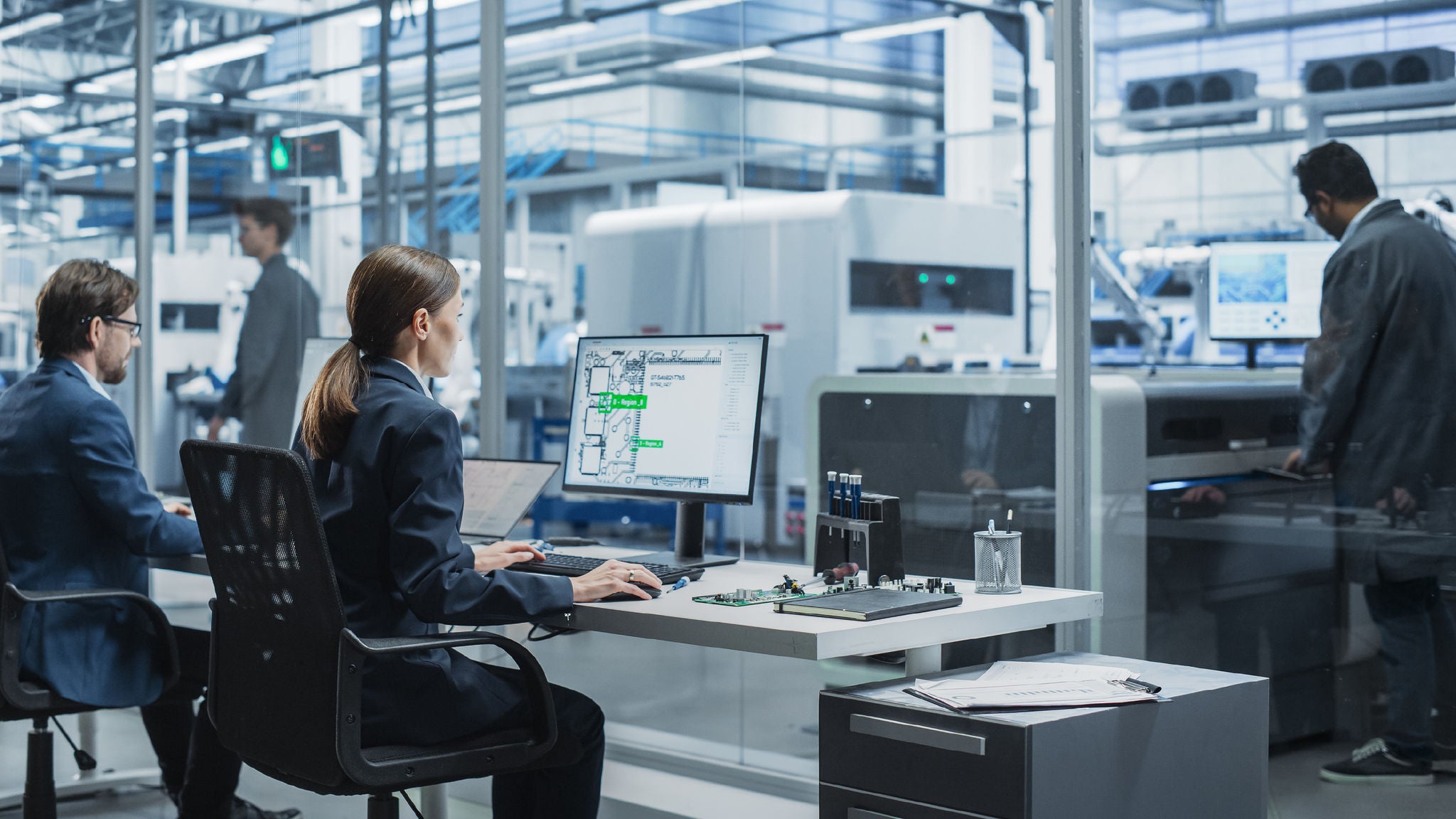 Engineers in a high-tech lab. A woman monitors CAD data on a desktop while others work around advanced machinery in a modern, glass-walled facility.