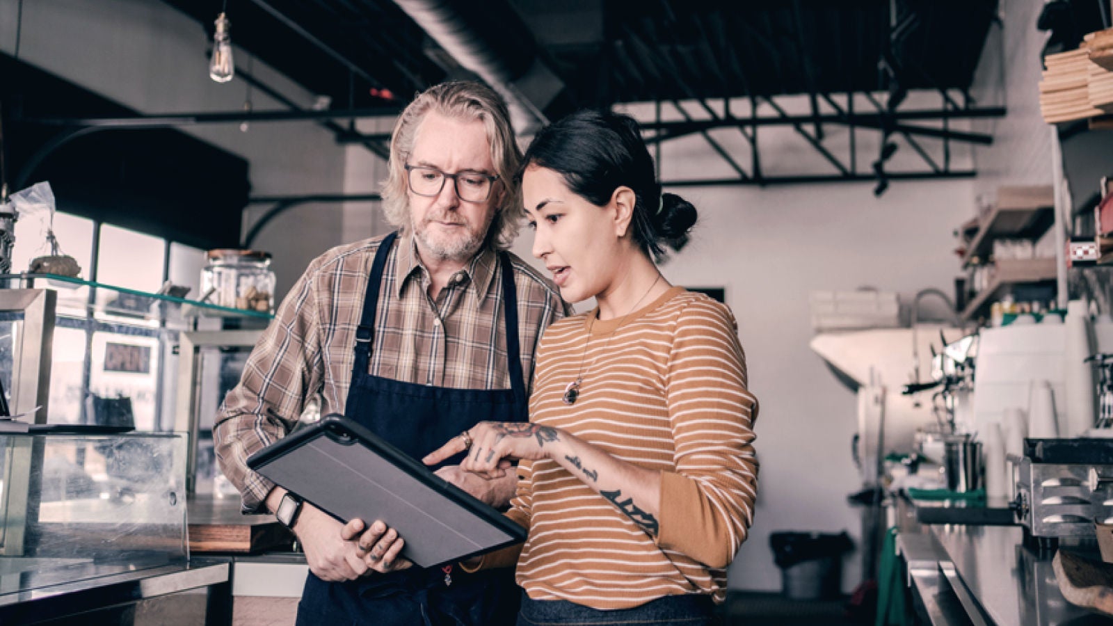 Two people standing inside a retail shop, looking at a tablet together while discussing plans. Shelves, counters, and store equipment are visible in the background.