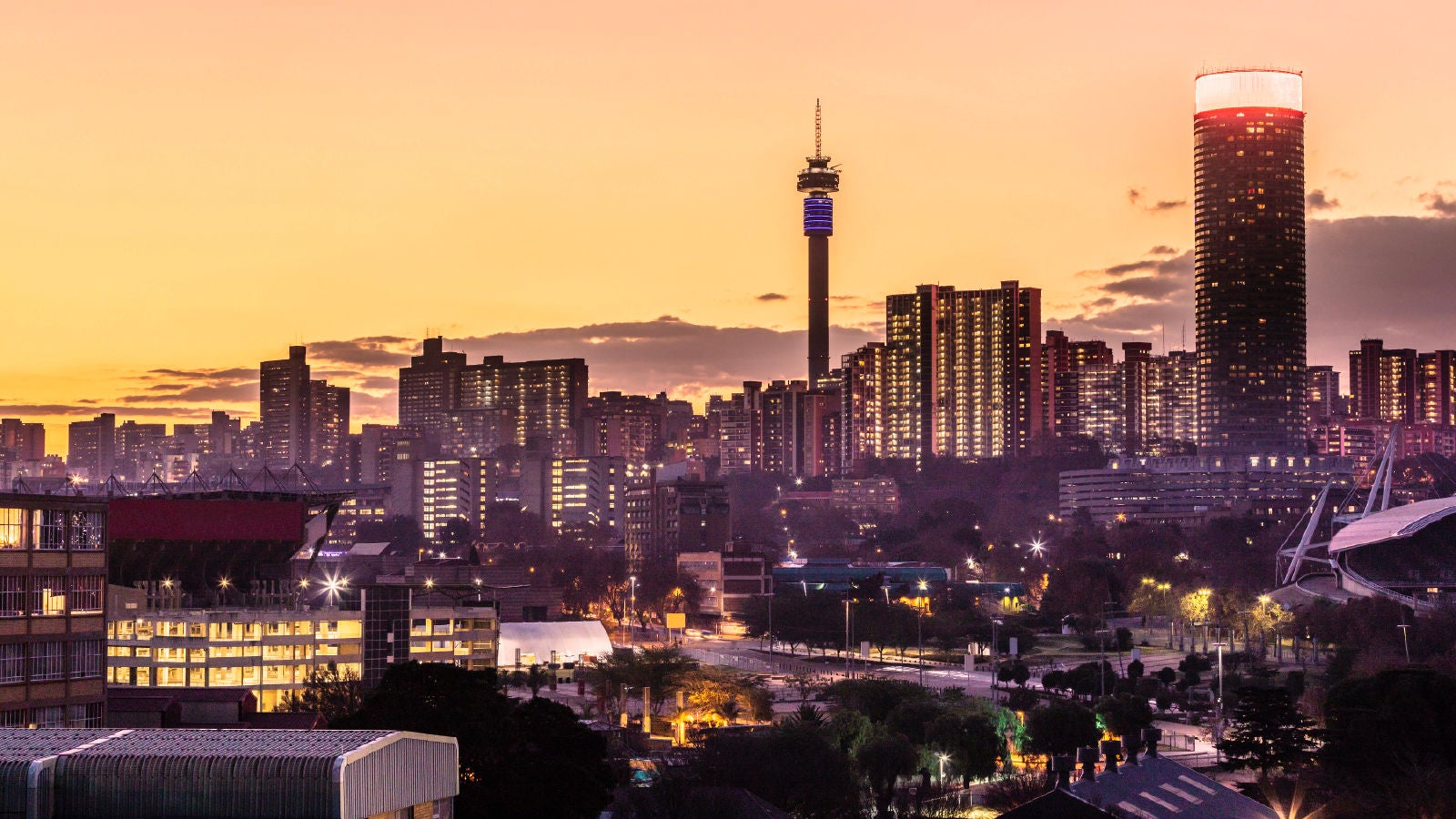 Johannesburg skyline at sunset with illuminated high-rise buildings, a communications tower, and city lights.